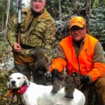 A proud hunter and his guide pose with two bagged ruffed grouse and a white bird dog.