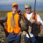 Two hunters with shotguns with one holding a harvested pheasant toward the camera.