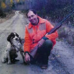 Bird hunter kneeling next to a bird dog holding a ruffed grouse in its mouth.