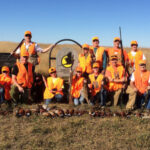 Large group of kids and adult bird hunters wearing orange vests pose by harvested pheasants.