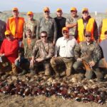 Large group of bird hunters posing behind a large pile of harvested pheasants.