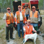 Five orange clad bird hunters with a pointing bird dog in front of a hunting rig.