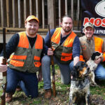 Bird hunters kneeling with bird dogs outside hunting lodge.