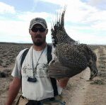 Bird hunter holding a harvested sage grouse toward camera with vast sagebrush landscape in background.