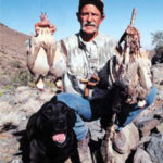 Bird hunter holding up harvested chukars kneeling by a black Labrador retriever.