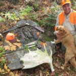 A bird hunter and dog next to a display of harvested ruffed grouse on a large flat rock adorned with a shotgun and moose antler.