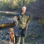 Bird hunter holding grouse next to dog.