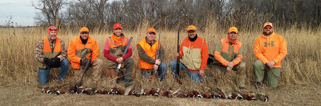 Seven bird hunters posing between field of prairie grass and a row of harvested rooster pheasants.