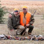 Two men kneeling behind row of harvested pheasants.