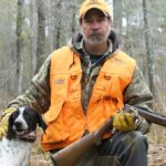 Kneeling hunter with shotgun petting bird dog.