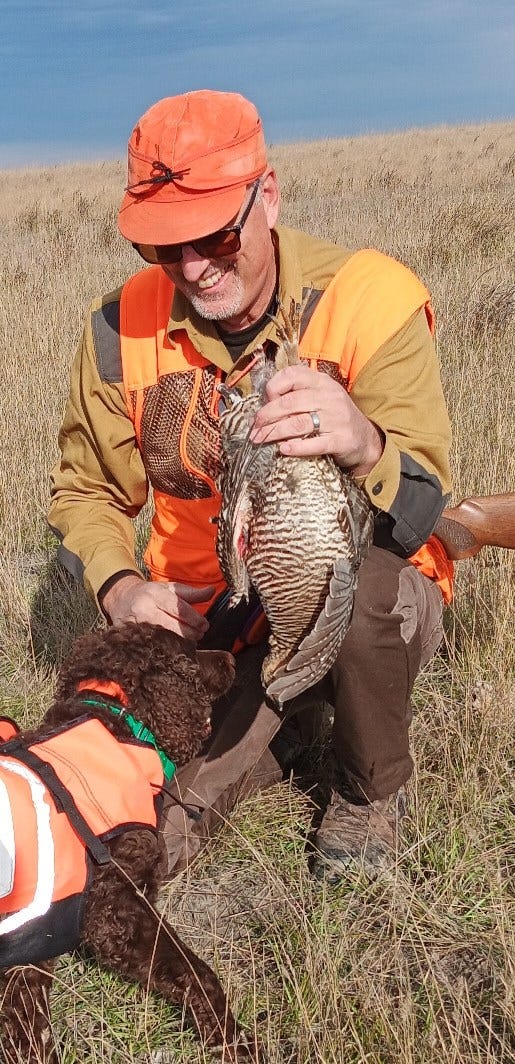 A smiling bird hunter accepts a retrieved prairie chicken during a successful hunt from a bird dog he trained himself.